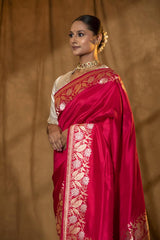 A woman modeling a magenta-pink Banarasi silk saree with gold and silver zari floral border, along with a matching running blouse piece.