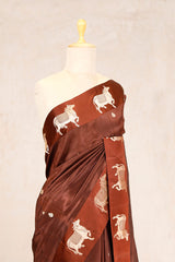 A mocha brown Banarasi silk saree with golden zari Pichwai motifs displayed on a saree rack, with a matching brocade design on the pallu and an unstitched blouse piece.