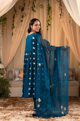 A woman in a blue traditional frock suit with a matching dupatta, standing in a decorated indoor setting.