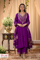 Woman in a purple traditional outfit standing in a decorated room with flowers and a table.