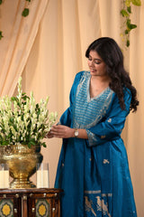Woman in a traditional blue salwar Suit holding a plant in front of beige curtains