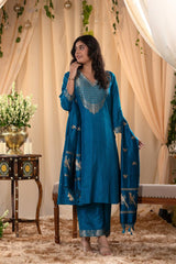 A woman in a traditional blue salwar suit and blue banarasi dupatta is standing in a decorated room.