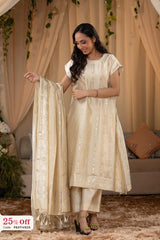 Woman in a beige traditional outfit with a dupatta, standing indoors with decorative curtains and plants.