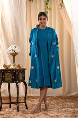 A woman in a blue Indo-Western dress with a matching coat is standing in front of beige curtains.