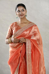 A woman modeling an orange tissue silk saree with silver zari woven stripes, paired with a contrast Banarasi silk semi-stitched blouse piece, giving a regal wedding or festive look