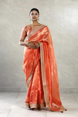 A woman modeling an orange tissue silk saree with silver zari woven stripes, paired with a contrast Banarasi silk semi-stitched blouse piece, giving a regal wedding or festive look