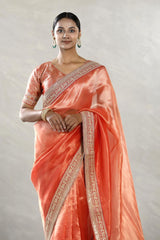 A woman modeling an orange tissue silk saree with silver zari woven stripes, paired with a contrast Banarasi silk semi-stitched blouse piece, giving a regal wedding or festive look
