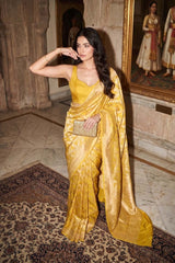 Woman draped in a Yellow Brocade Banarasi Silk Saree featuring an intricate gold zari woven ethnic floral jaal pattern, holding a gold clutch in a heritage setting.