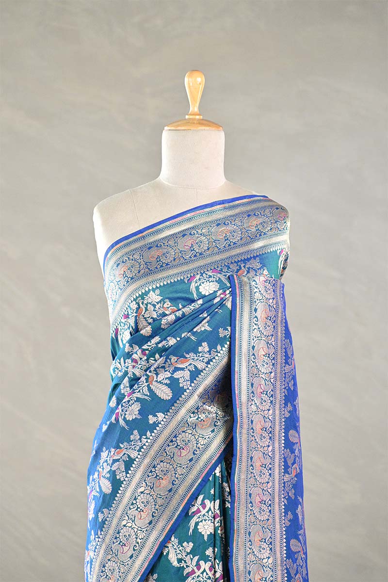 Teal blue Banaarsi silk saree with floral and meenakari zari work draped on a mannequin against a gray background