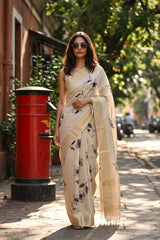 Woman in a beige saree with floral patterns standing next to a red postbox on a street.