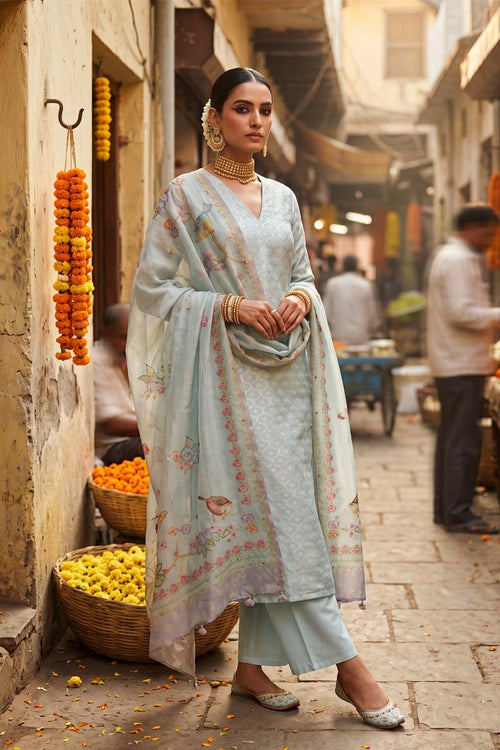 A woman stands wearing a light pastel blue traditional outfit with a matching dupatta, decorated with delicate floral embroidery.