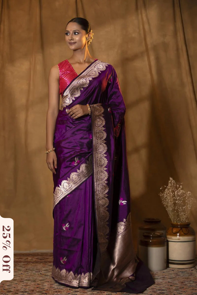 Woman wearing a purple saree with silver border against a beige curtain background