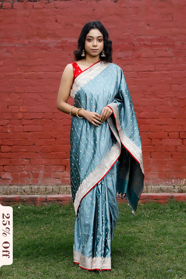 Woman in a blue saree with white and red border standing against a red brick wall.