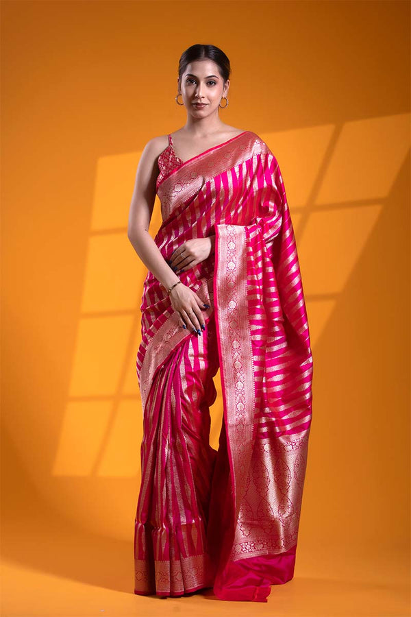 A woman modeling a rani pink handloom katan silk Banarasi saree with gold zari floral stripes, along with a matching running blouse piece.