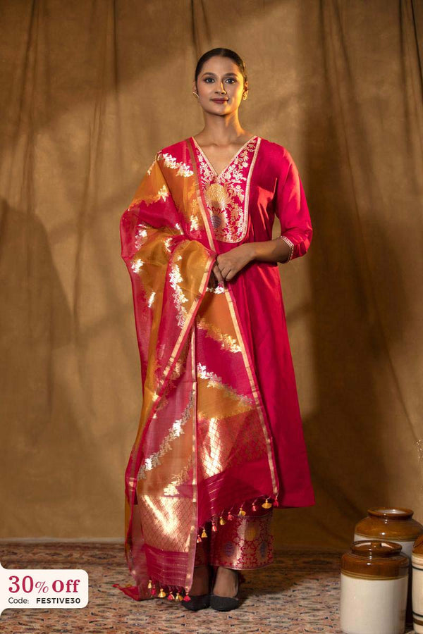 Woman in a red saree with gold patterns standing against a brown curtain.