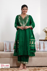 Woman wearing a green traditional outfit with gold embroidery in a living room setting.
