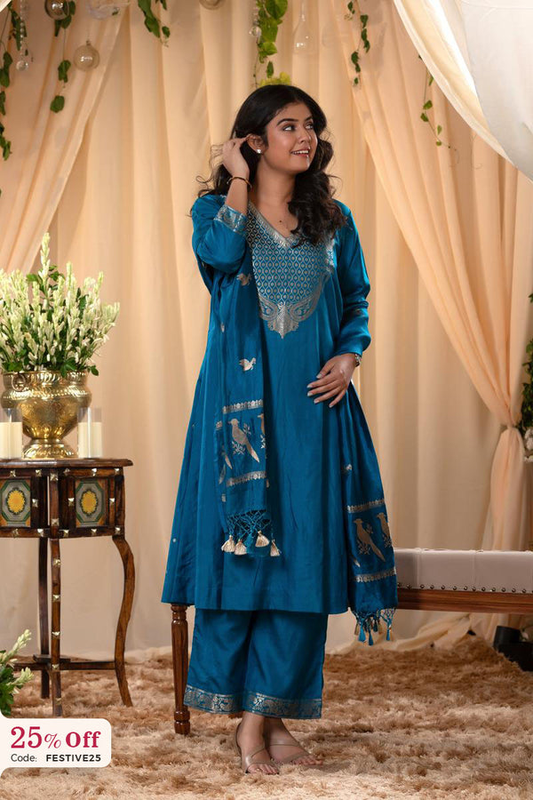 Woman in a blue traditional outfit with a decorative scarf, standing in a decorated room.