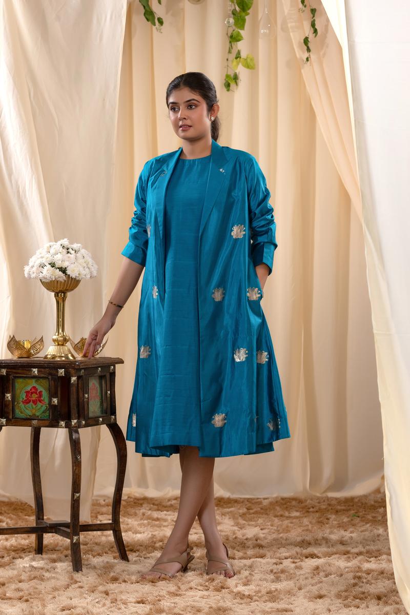 Woman in a blue Indo-Western outfit standing next to a small table with decorative items against a beige curtain background.
