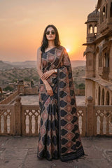 Woman in a patterned tussar silk saree standing on a stone platform with a sunset and architectural structure in the background.